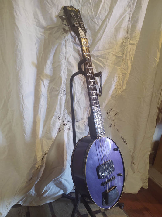 A custom Bishop Blueridge Tinjo Bass with a purple-to-blue shifting finish, 4-string setup, dual pickups, and a resonator-style metal body, displayed on a stand against a white fabric backdrop.
